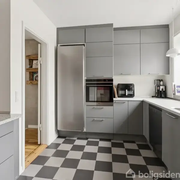 Gråt køkken med indbyggede skabe, køleskab og ovn. Rum med ternet sort-hvidt gulv og store vinduer med lysindfald. En kaffemaskine står ved siden af vasken.