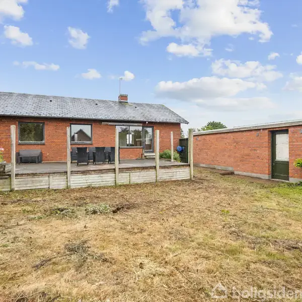 Rødt murstenshus med terrasse, udstyret med havemøbler, omgivet af en græsplæne og med en tilstødende garage. Himlen er delvist skyet, hvilket skaber en typisk dansk havescene.