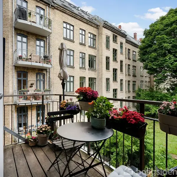 En balkon har et rundt bord med blomsterpotter, omgivet af blomstrende planter. Bygninger i mursten kan ses i baggrunden, samt træer der omfavner gårdspladsen.