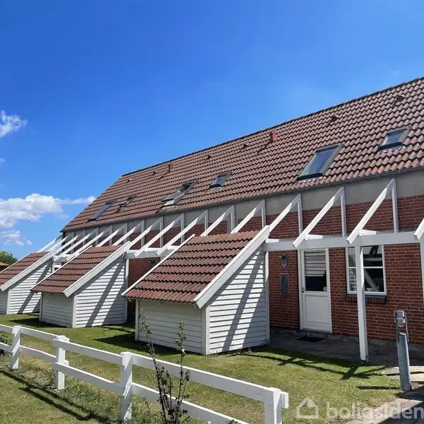 Baggård med rækkehus; skråt tag, røde teglsten, hvidmalet trækonstruktioner. Grøn græsplæne med et hvidt hegn i forgrunden under blå himmel med spredte skyer.