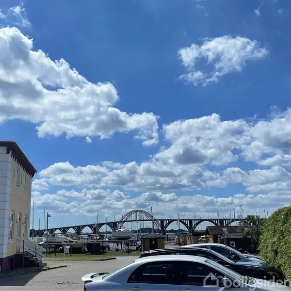 Bygning står stille ved siden af parkerede biler på en solrig dag. En buet bro strækker sig over jernbanespor i baggrunden med en klar blå himmel og spredte skyer.