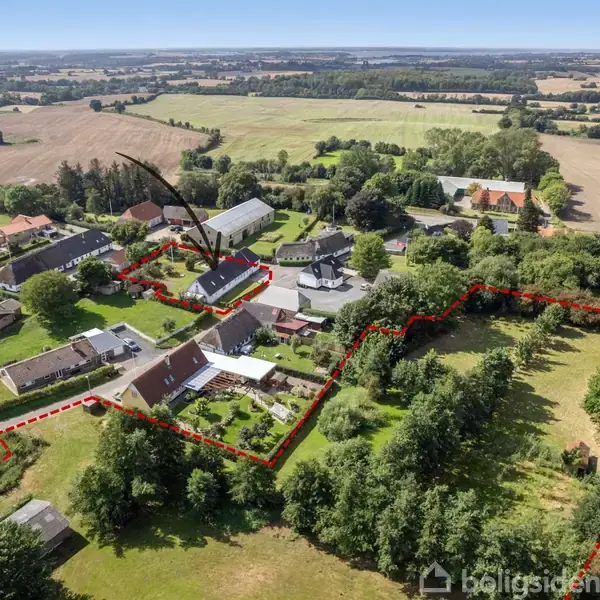 Et hus er markeret med en rød prik og pile omkranset af områder med marker og træer. Flere huse og grønne områder i landsbyen er synlige omkring det.
