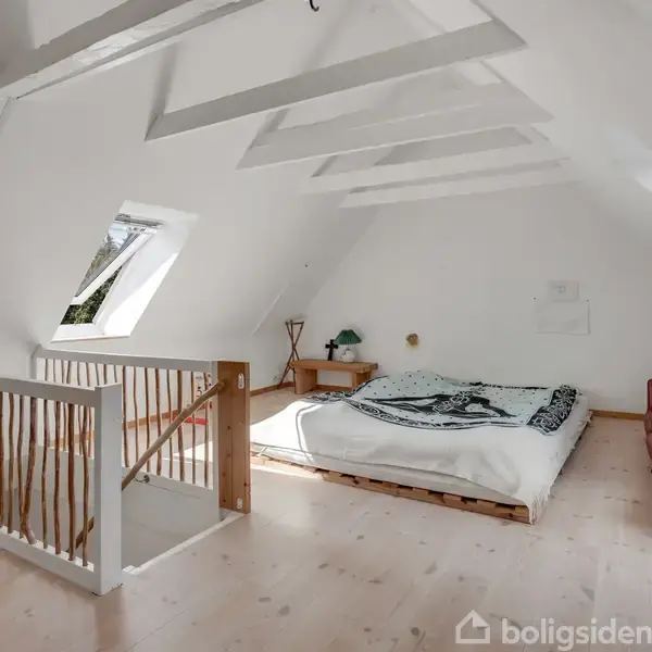 Et lyst rum på en loftsetage med en seng centralt placeret. En trappe leder op til rummet, som har skrå vægge og vinduer. Møbler inkluderer en kommode og en sofa.