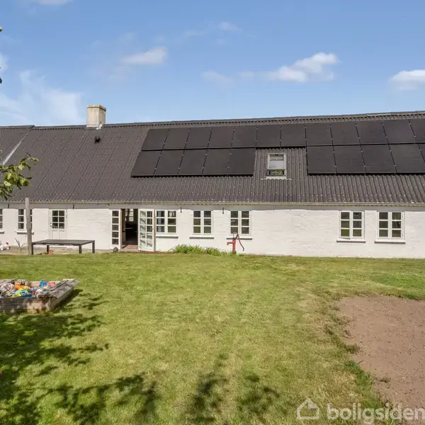 Hvidmalet hus med solpaneler på taget. Det står stille mellem en grøn have og en sandkasse i græsset. Skyfri himmel og spredte træer omkring. Tekst: "boligsiden".
