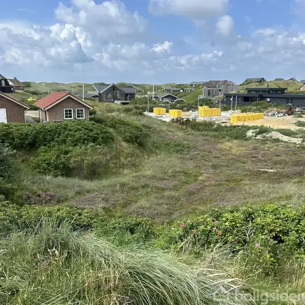 Feriehuse omkranser græsbevokset sandet klitlandskab, hvor bygninger har røde og mørke tage. En byggeplads med gule materialer ses til højre under en overskyet himmel.