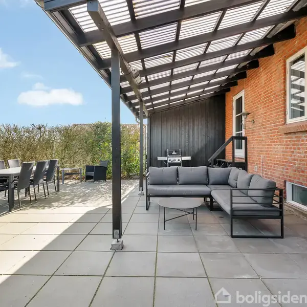 Terrasse med grå sofa under en delvis overdækning ved siden af et rødt murstenshus. Spisebord med stole står i solen. Grønne buske omkranser området.