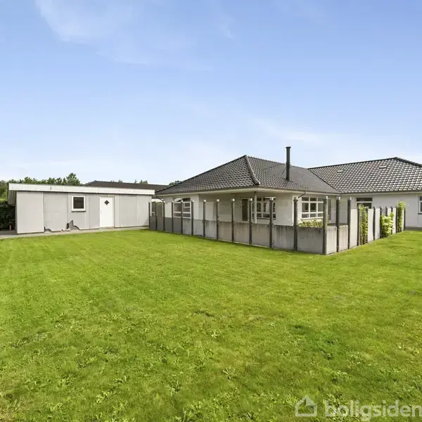 Et hvidt hus med sort tag står i en velplejet have omkranset af hegn. Græsset er frodigt, mens himlen er blå og skyfri.