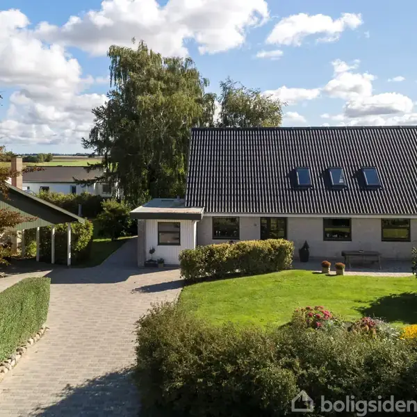 Hus med sort tag og tre ovenlysvinduer, omgivet af velplejet have med buske og græsplæne. Stenbelagt indkørsel fører op til huset under en solrig himmel med få skyer.