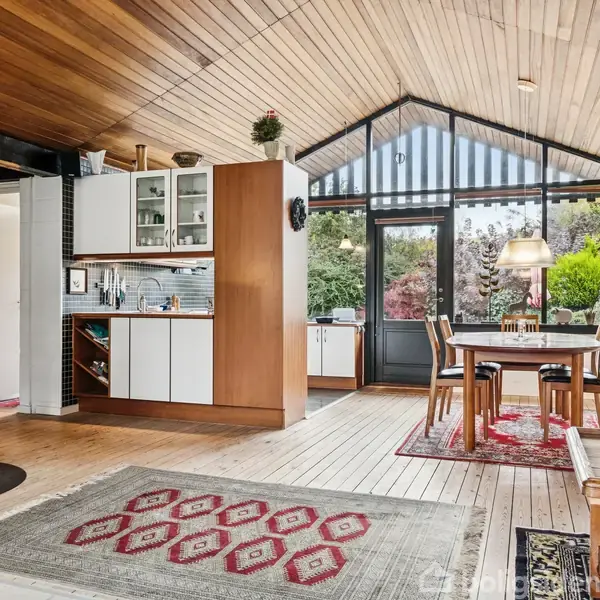 Brændeovn opvarmer stue med trægulv, hvor spisebord står. Køkken ligger til venstre. Store vinduer viser frodig have udenfor. Rummets loft er beklædt med træpaneler.