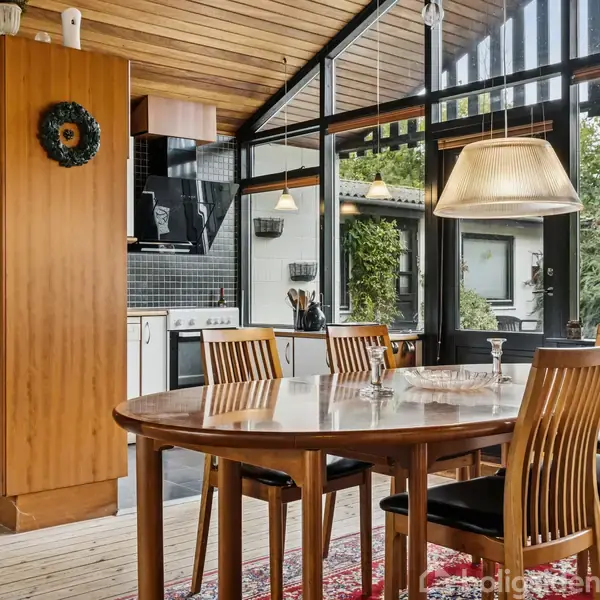 Et rundt spisebord i træ står i en solrig spisestue med fem stole omkring det. Rummet har store vinduer, der viser en grøn have udenfor. En stor lampe hænger over bordet.