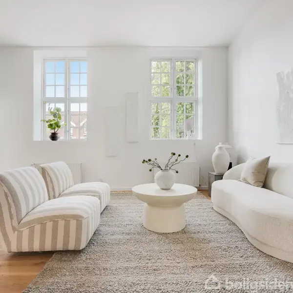En moderne stue indeholder en hvid sofa, stribede stole og et rundt bord på et beige tæppe. Værelset er lyst med store vinduer og minimalistisk kunst på væggen.