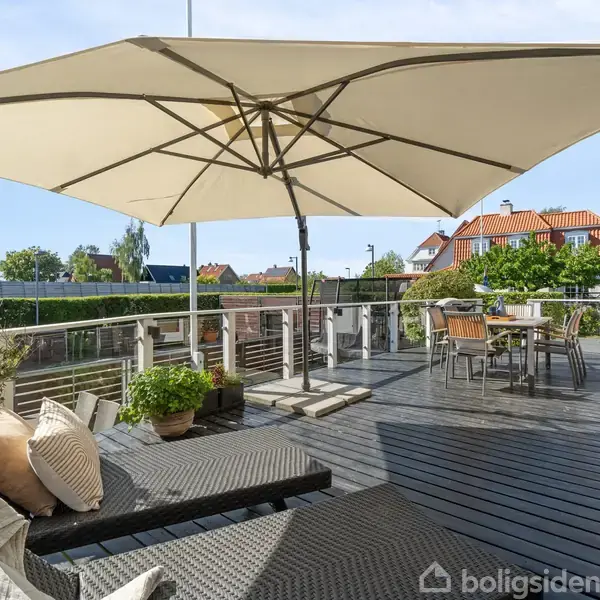 Stor parasol dækker terrasse med liggestole og spisebord i et moderne udendørsområde. Omkring ses planter og et glimt af et boligkvarter med huse og blå himmel.