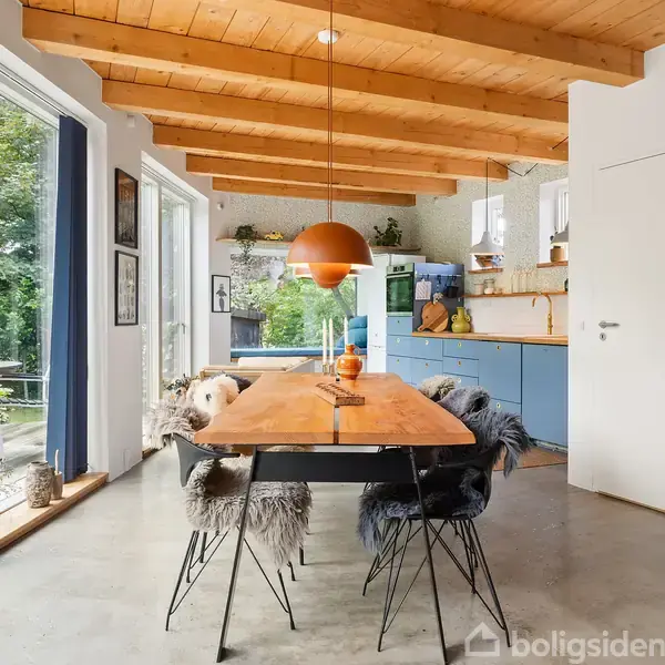 Træbord med fåreskindstole står under en orange lampe i en moderne spisestue. Rummet har åbent køkken, lyse vægge, træloft og store vinduer med udsigt til terrasse og grønne omgivelser.