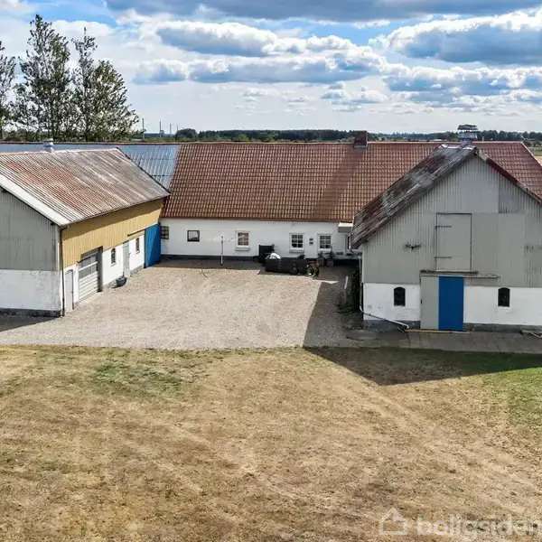 To bygninger med røde tegltage står på en gårdsplads omgivet af åbne marker og træer under en blå himmel med skyer.