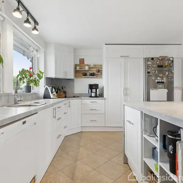 Køkken med hvide skabe og bordplader, kaffemaskine på bænken. Køleskab med magneter til højre. Flisegulv og kogeplade ved det store vindue med planter, som leder dagslys ind.