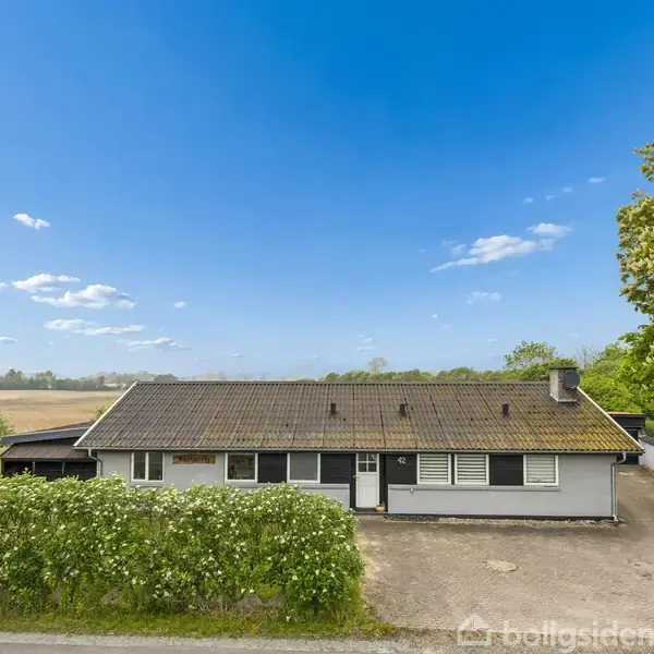 Hus med fladt tag og carport til venstre, beliggende ved en vej. Foran huset er en lav busk, og omgivelserne består af åbne marker og træer under en klar blå himmel.