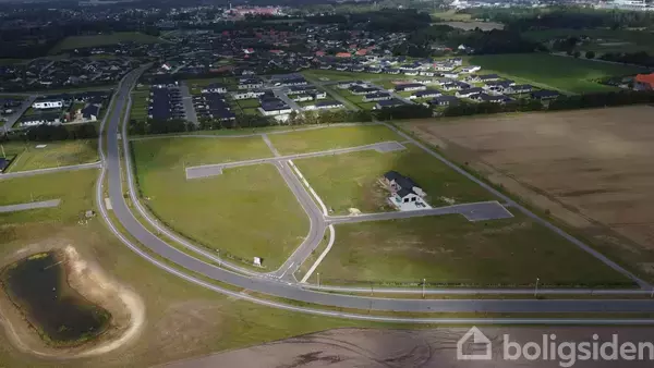 Et nybygget hus står på en stor åben grund. Omkring ligger veje og marker, med en by i det fjerne.