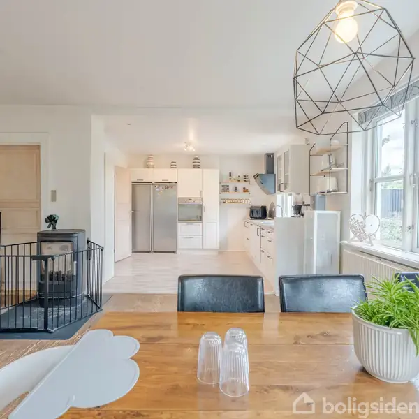 Et spisebord med en plante og drikkeglas står centralt i rummet. Et moderne køkken med hvide skabe findes i baggrunden, ved siden af store vinduer, som giver naturligt lys.