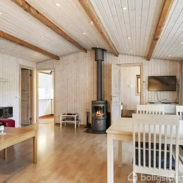 En stue har et brændeovn i drift, skaber hygge. Omkring er røde sofaer, træbord, spisebord med stole og vægmonteret TV. Værelset har lyse trævægge og et lyst trægulv.