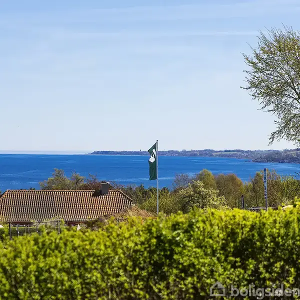 Flag vimpler vejrer nær et rødt tegltag med udsigt til havet i baggrunden; omgivet af frodigt grønt landskab og en stor træ.