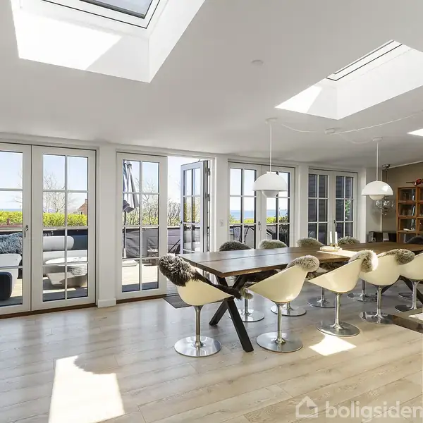 Stue med spisebord omgivet af seks stole og lodretstående reoler. Store vinduer og ovenlysvinduer giver rummet naturligt lys. En lædersofa står til venstre, og døre åbner ud til terrassen.