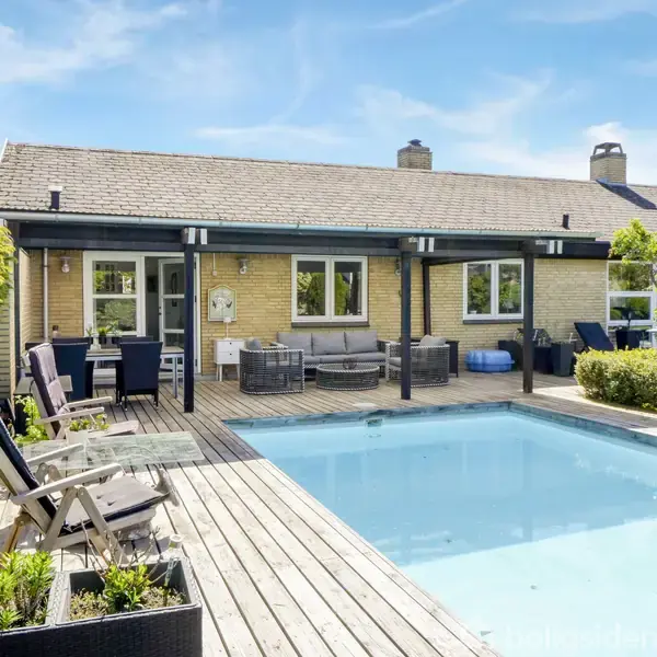 En swimmingpool omgivet af et træterrasseområde med solstole og planter, foran en gul murstensvilla med store vinduer og havemøbler under en overdækket terrasse.
