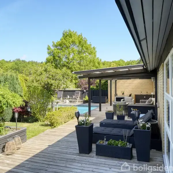 En træterrasse med moderne havemøbler og planter. Ved siden af er en have med grønne buske og træer. I baggrunden ses en overdækket terrasse og poolområde.