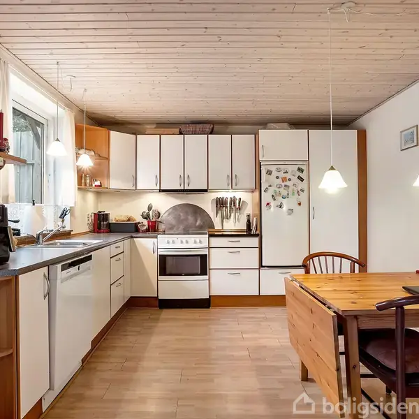 Et funktionelt køkken med hvide skabe og træelementer. Har opvaskemaskine, ovn og køleskab. Ved siden af står et spisebord med stole. Køkkenet er lyst med to hængende lamper.