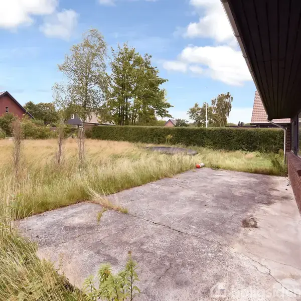 En betonterrasse med nogle pletter strækker sig langs siden af et murstenshus. I nærheden vokser højt græs, og nogle træer og buske kan ses i baggrunden under en blå himmel.