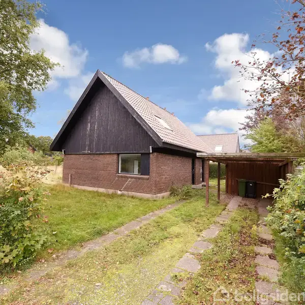 Et brunmurstenshus med sort tag ligger ved siden af en grussti omgivet af frodigt grønt. En carport ligger ved siden af huset, og der er et postkasse på venstre side.
