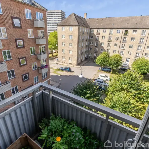 Balkon med grøn plante i forgrund; udsigt over gård med træer, parkerede biler og murstensbygninger. Skilt med teksten: "Ny bolig? 70 60 27 00" ses til venstre.