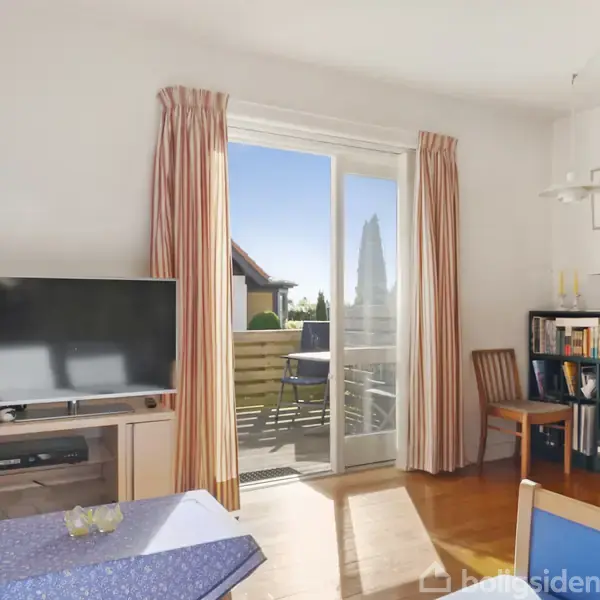 TV står på et lavt bord og viser en sort skærm. Ved siden af er en åben skydedør med adgang til en balkon. Reol med bøger indikerer et hjemligt rum.