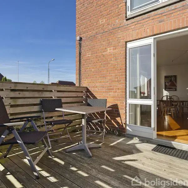 Terrasse med to stole og et bord, åbne døre til en lys stue. Omgivet af et træhegn, teglstensmur med klar blå himmel i baggrunden.