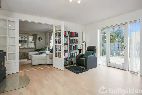 En stue med en reol fyldt med bøger mellem to lænestole, der står tæt ved en glasdør til en terrasse. Til venstre er en sofa og bord i et tilstødende rum.