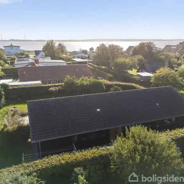 Et hus ligger i en grøn have med hække, omgivet af flere huse. I baggrunden kan man se en sø eller fjord.