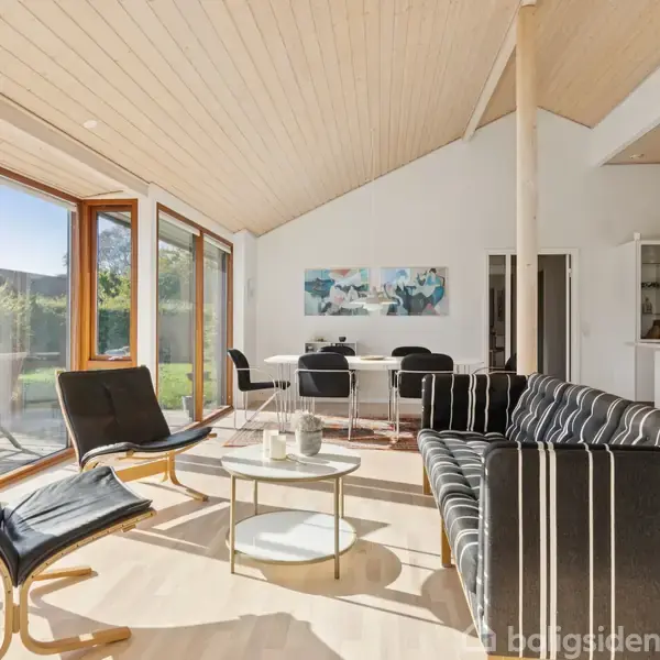 En stue med stribet sofa og sorte stole omkring et lavt bord. Store vinduer slipper sollys ind, mens en spiseplads med kunst på væggen ses i baggrunden.