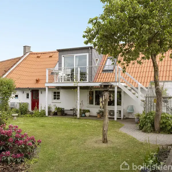 Et hus med rød tegltag har en balkon med glasrækværk og udendørs trappe. Omgivet af en velplejet have med blomster og træer.