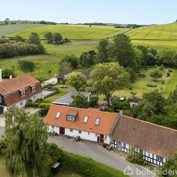 Et bindingsværkshus med orange tag ligger ved en landevej, omgivet af frodige marker og grønne bakker. Flere træer og naboens huse skaber en idyllisk landskabsramme.