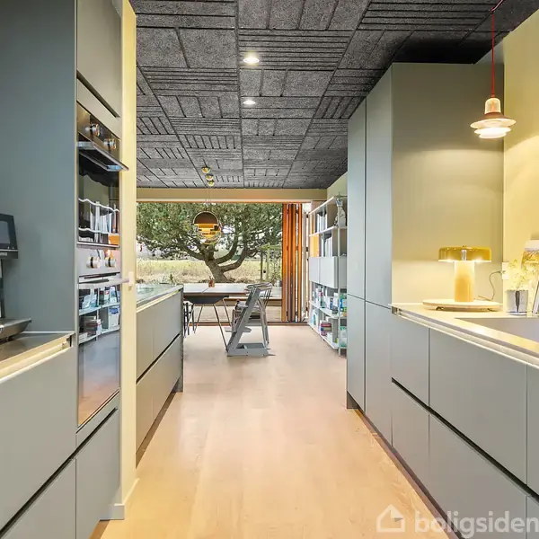 Køkken med moderne grå skabe strækker sig langs begge sider. Kaffemaskine og vask er synlige. Spisebord ses for enden af rummet med udsigt til træ og vindue.