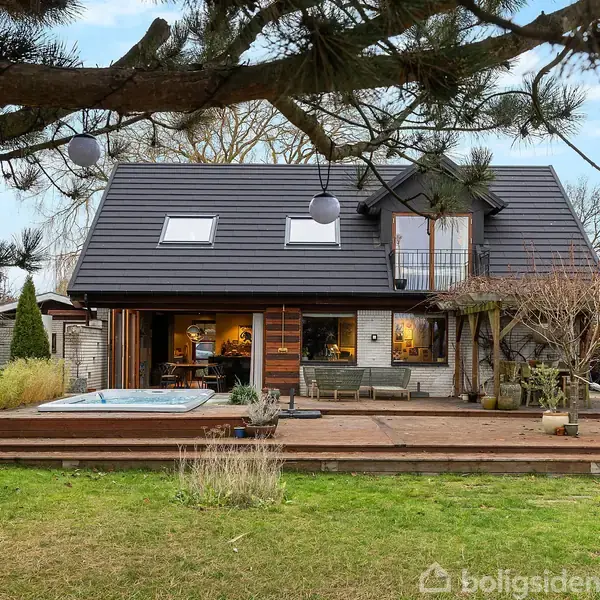 Et moderne hus med sort tag ses fra haven. Foran er en træterrasse med en pool og hængekøje, omkranset af træer og buske i en hyggelig udendørs atmosfære.