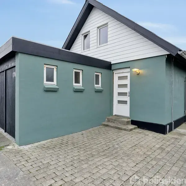 Huset har blågrønne vægge og en hvid hoveddør med trin. Det har flere små vinduer og ligger på en brostensbelagt gårdsplads under en blå himmel.
