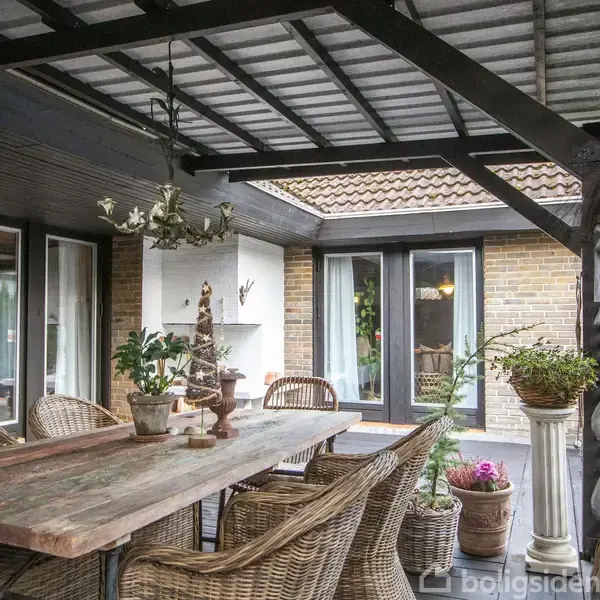 Et rustikt spisebord omgivet af flettede stole står på en overdækket terrasse. Planter i potter fylder rummet, og store vinduer åbner op til husets indre.