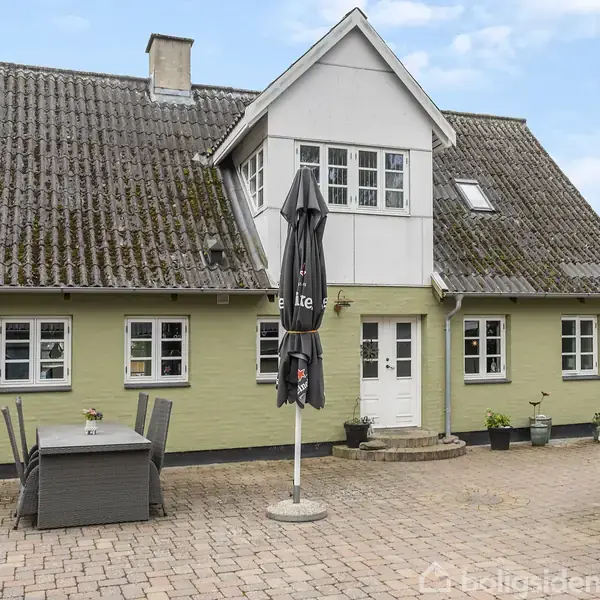 Gult, klassisk hus med skråt tag og kvist. Møbelopstilling og parasol på flisebelagt terrasse. Grill ved siden af, omringet af grønne planter. Ingen synlig tekst bortset fra “Lite” på parasollen.