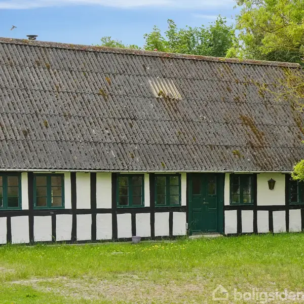Bindingsværkshus med sort-hvide mure og et mørkt tag. Huset står stille i en grøn have, omgivet af træer og blå himmel. En grøn dør er centralt placeret.