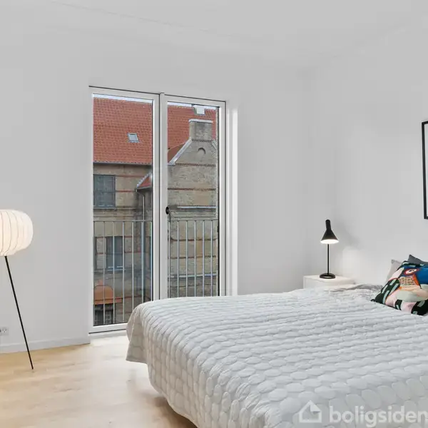 Et soveværelse med en stor seng dækket af et hvidt tæppe. Ved siden af står en moderne lampe, og der hænger en billedramme på væggen. Rummets vindue viser udsigt til en bygning med rødt tag.