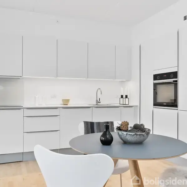 Et moderne køkken med hvide skabe. Bordet har en rund trætoppet flade med dekorative genstande. I baggrunden ses en indbygget ovn og køleskab. Rummet er lyst og minimalistisk.