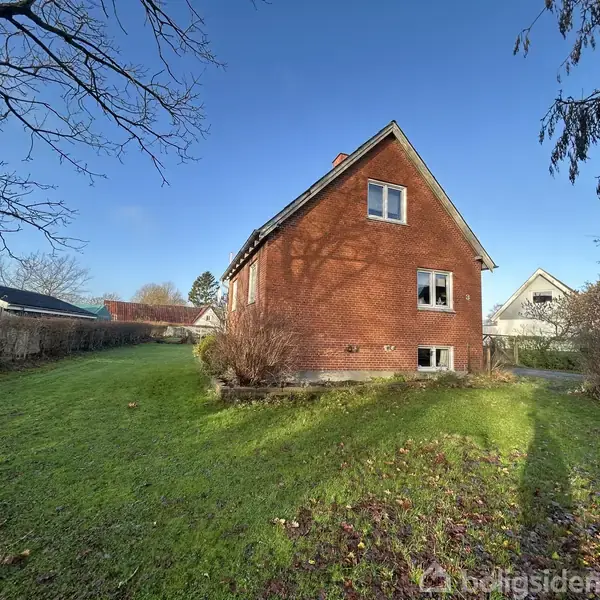 Rødt murstenshus står på en grøn græsplæne omgivet af buske og mindre træer under en klar blå himmel, omgivet af andre huse i et boligkvarter.