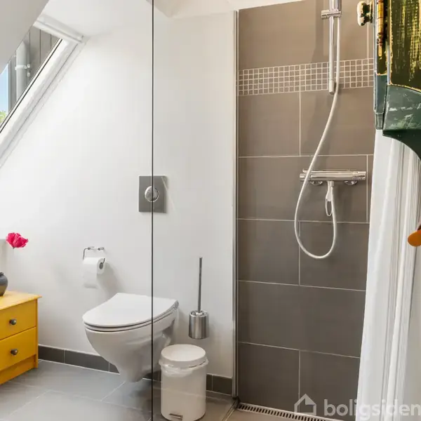 Badeværelse med brusekabine og grå fliser. En monteret toiletskål ved siden af en gul kommode med en vase. En træabe hænger på en hylde. Sandfarvet gulv.