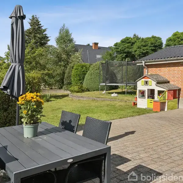 Terrassebord med stoler og parasoll står på fliser. En potte med gule blomster er på bordet. I baggrunden ses en have med et gyngestativ og en lille legehytte ved et rødt murstenshus.