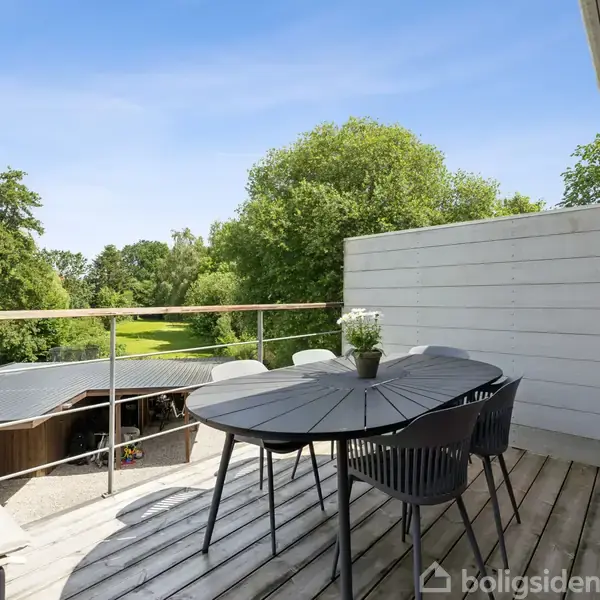 Ovalt bord med sorte stole står på en terrasse af træ. En plante pynter på bordet. Udsigt over grøn have med træer i baggrunden under en blå himmel.